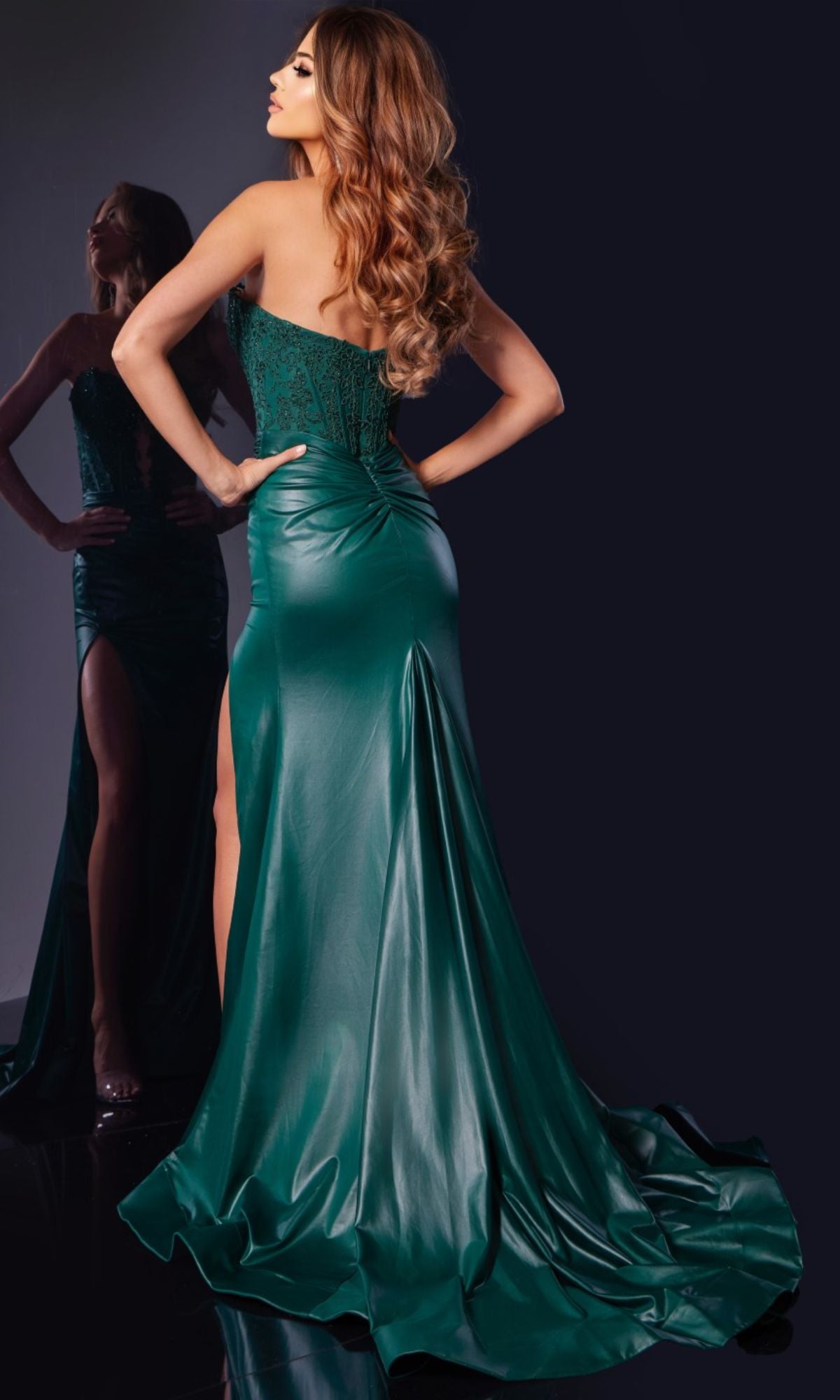 A woman poses with her back to the camera in the Jovani 43267 Strapless Tight Long Prom Dress, an emerald green, floor-length gown with a high slit; her reflection shines in the mirror as she glances over her shoulder.